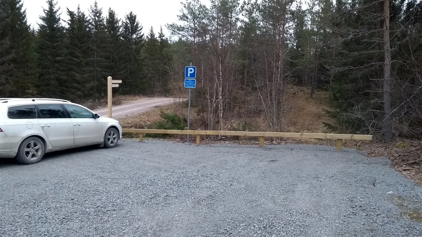 En bil står på grusad parkeringsplats vid en skogsbilväg. Runt parkeringen finns en upphöjd kant och vid sidan står en vägvisare och parkeringsskylt.
