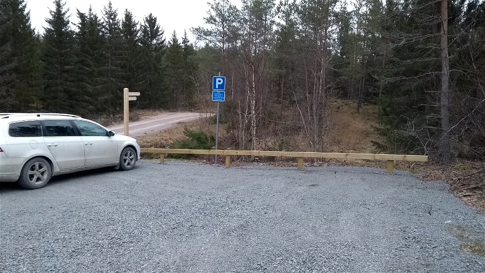 En bil står på grusad parkeringsplats vid en skogsbilväg. Runt parkeringen finns en upphöjd kant och vid sidan står en vägvisare och parkeringsskylt.
