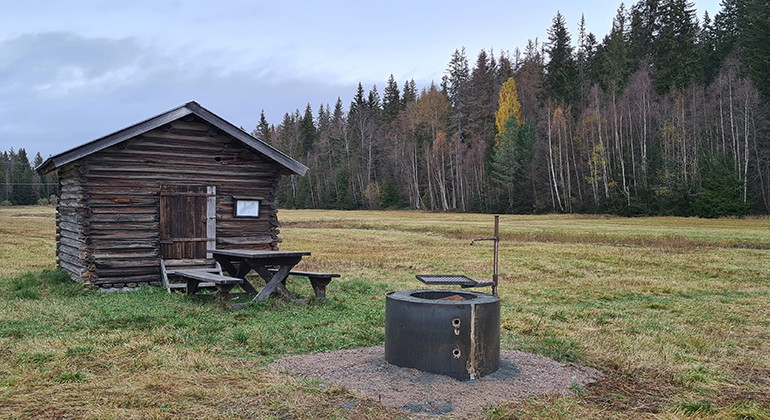 Ett litet hus på en äng med eldstad utanför. Foto.