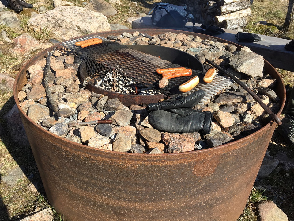 Rund grillstad av järn och sten. På grillen ligger korvar och korvbröd. bredvid grillen ligger ett par tumvantar i skinn.