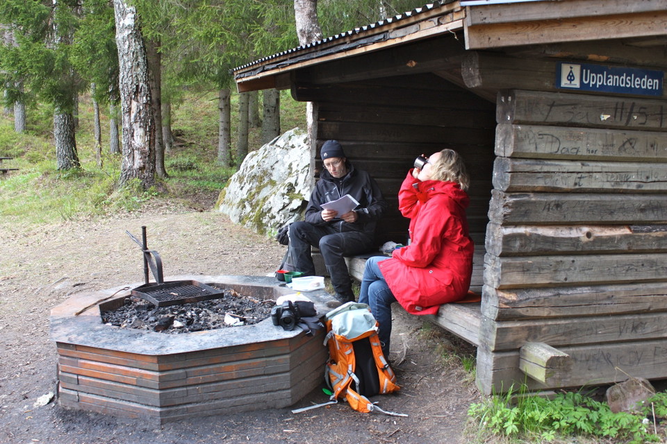 En man och en kvinna sitter i ett vindskydd i skogen. Framför vindskyddet står en grillplats. 