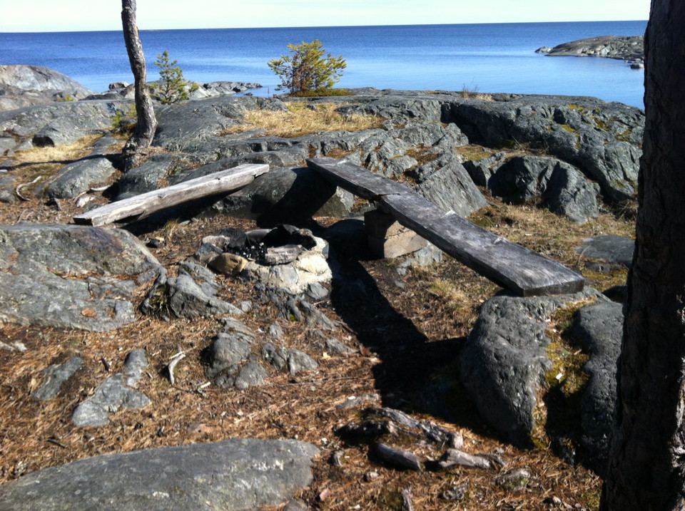 En låg eldstad med två stockbänkar ovanpå stora stenar. Det är väldigt ojämnt och stenigt på marken runtom grillplatsen.
