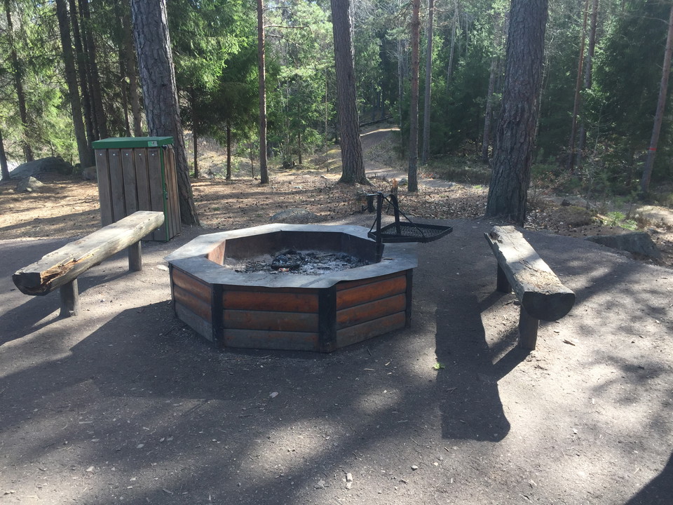 Grillplats med två sittbänkar och en soptunna i närheten. I bakgrunden är det skog.