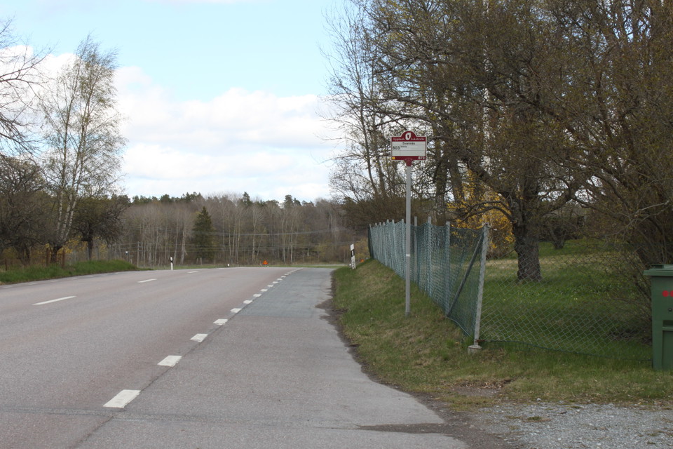 Vid en bilväg finns en ficka med skylt för busshållsplats. Vid skylten finns ett staket.