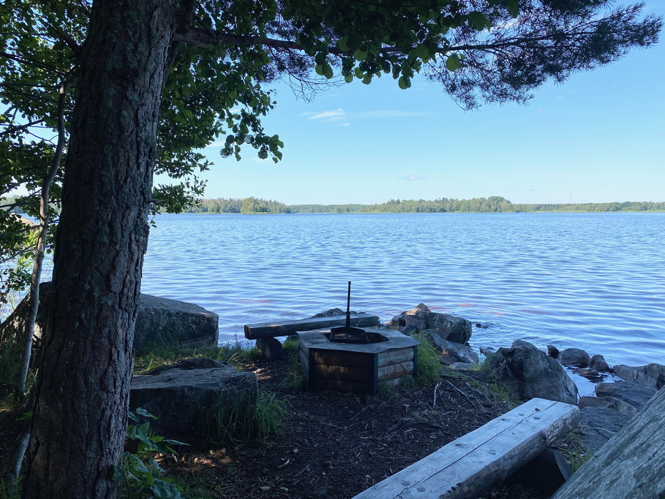 En grillplats vid en sjö. Runtom platsen finns flera stora stenblock och ett träd.