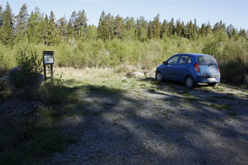 En bil står på en mycket liten grusad parkeringsplats vid utkanten av en skog. På platsen finns en informationstavla.