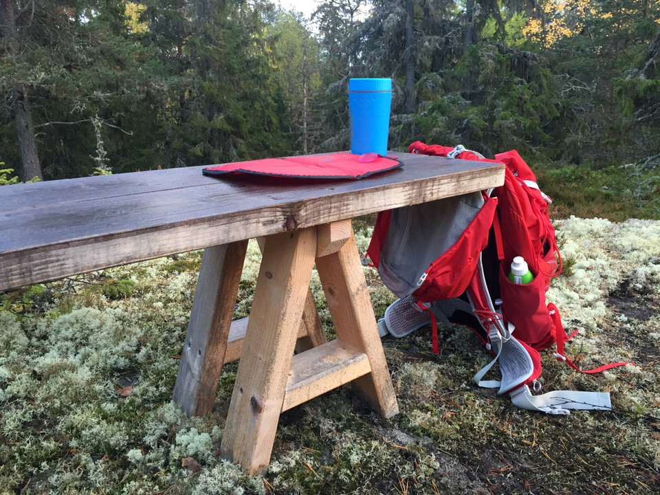 Ryggsäck, sittunderlag och mattermos på bänk vid rastplats.