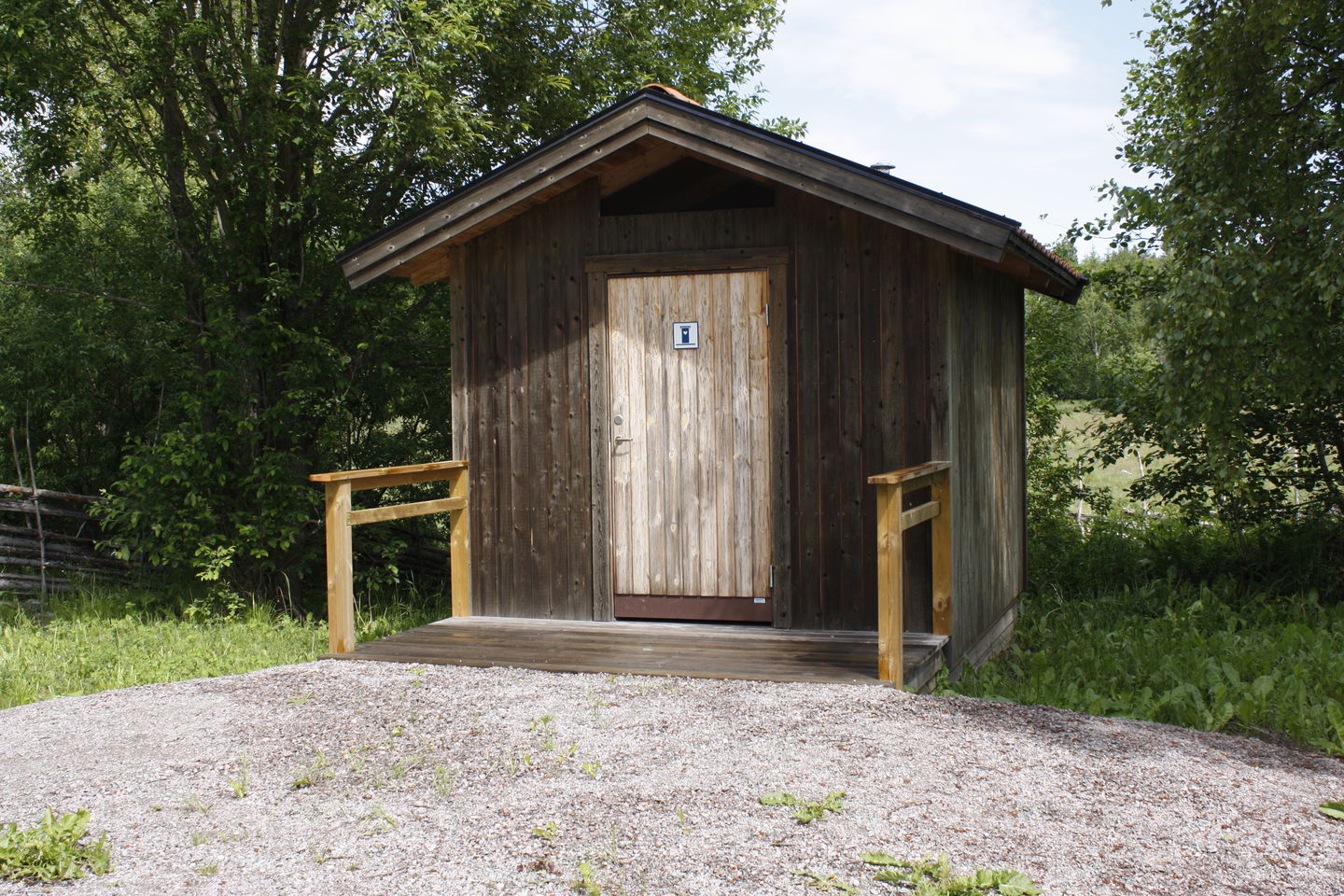 En bred ramp med handräcken leder fram till en torrtoalett. Framför rampen är det en grusplan.