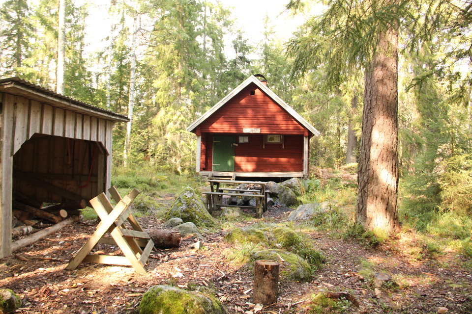 Vedförråd, bänkbord, eldstad. Stugan i bakgrunden.