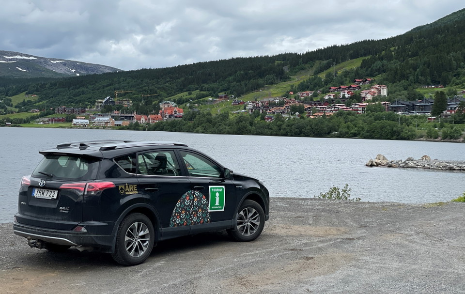 En svart bil med ett turistinformations I på en skylt på sidan ståendes vid en sjö med berg i bakgrunden