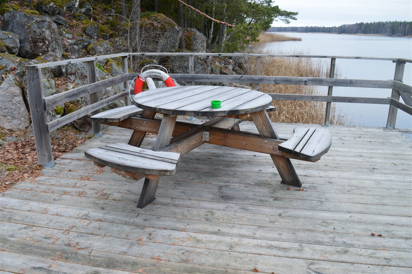 Närbild på ett bänkbord med fyra sittplatser som står på ett trädäck vid vattnet. Runt däcket finns ett högt skyddsräcke.