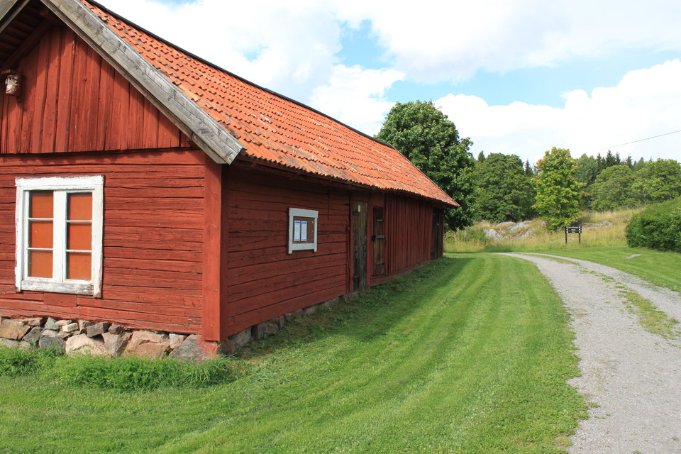 En äldre lada med fönster står på en gräsmatta vid en grusad bilväg.