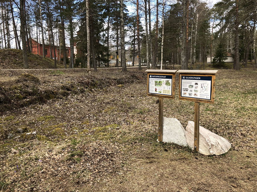 Två informationsskyltar. Röda byggnader långt i bakgrunden.