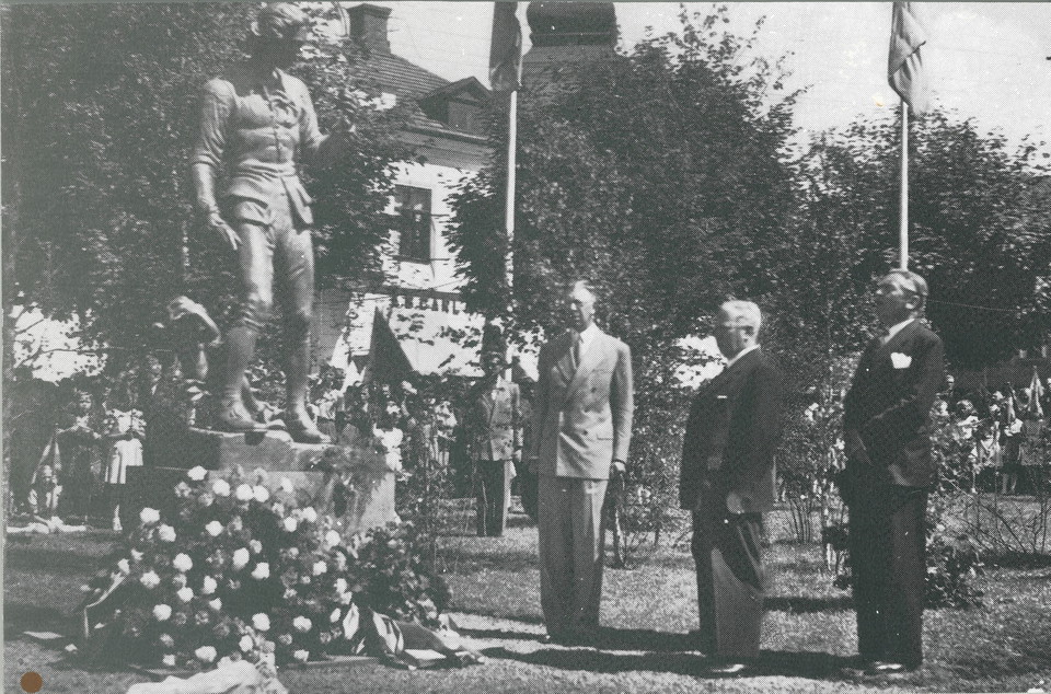 Tre män står på rad intill en staty. Vid foten på statyn ligger en stor blomsterkrans.