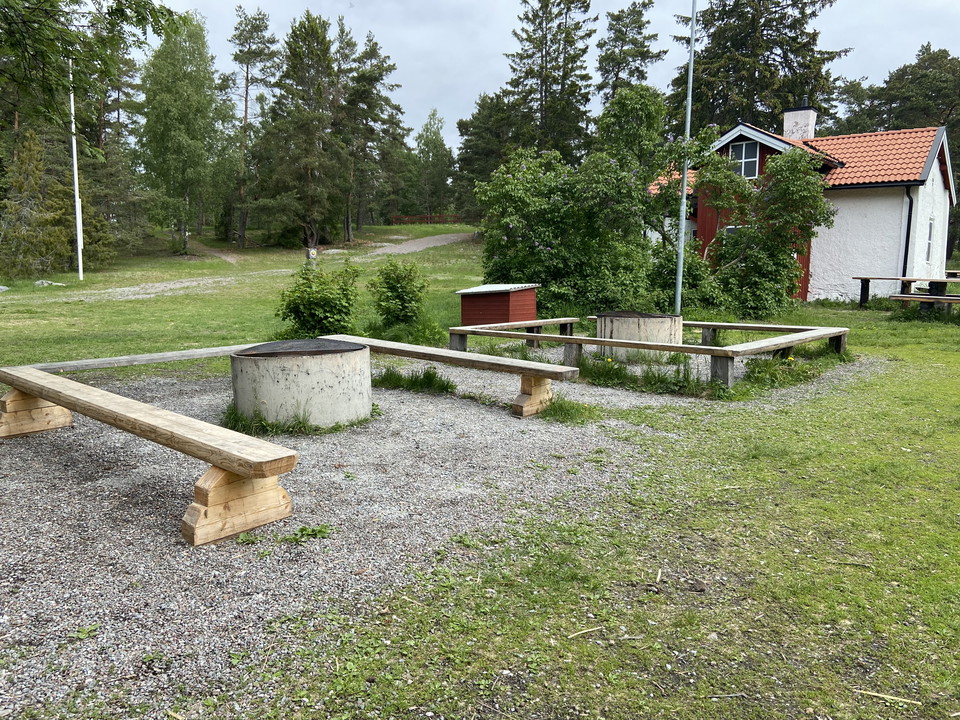 Två eldstäder i form av cementrör med galler och sittbänkar runt om. I bakgrunden syns en liten stuga.