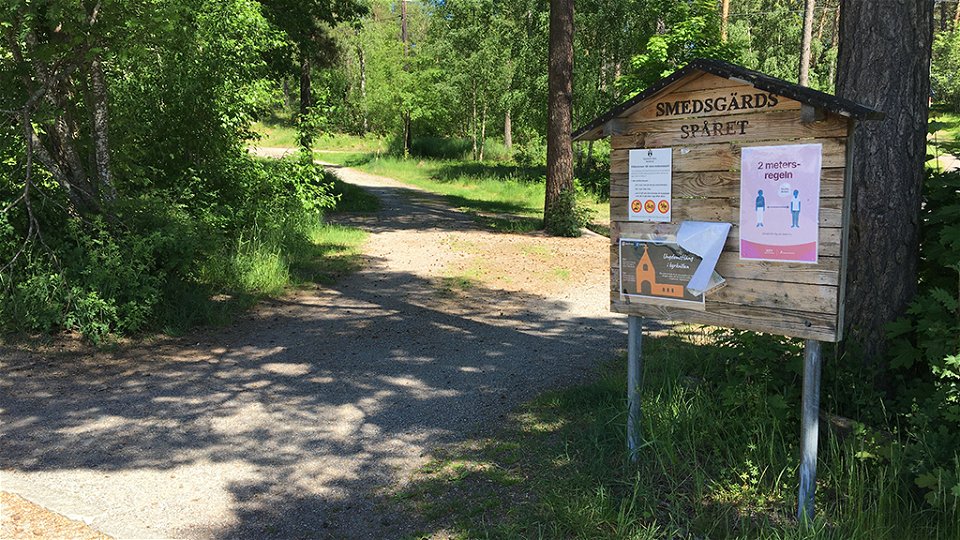 En träskylt med texten Smedsgärdsspåret och affischer. Skylten står vid en grusad stig i skogsbrynet.