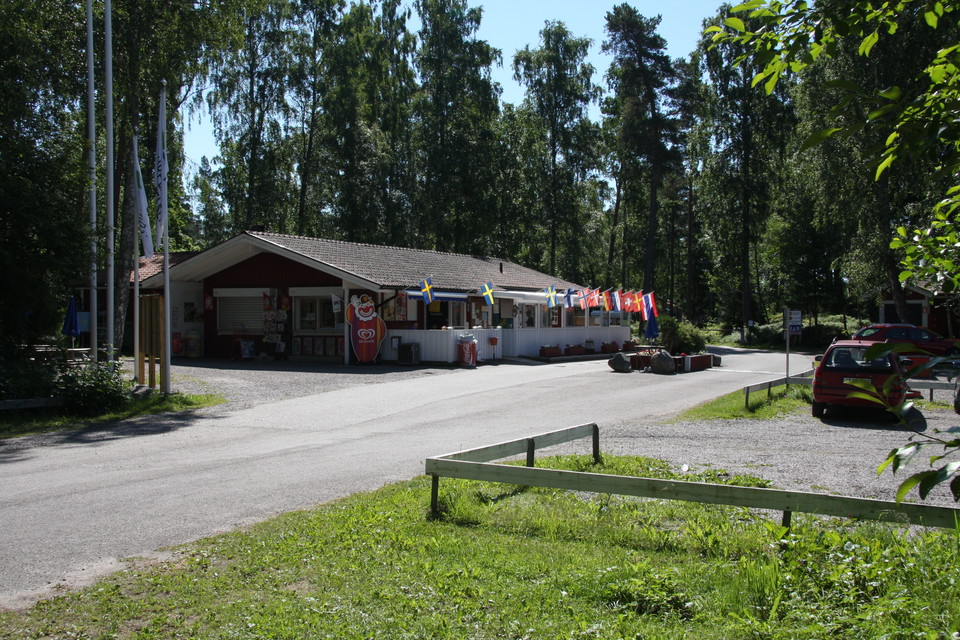 En asfalterad väg leder fram till campingbyggnaden. På andra sidan vägen finns en parkeringsplats med grus och lite gräs på marken.