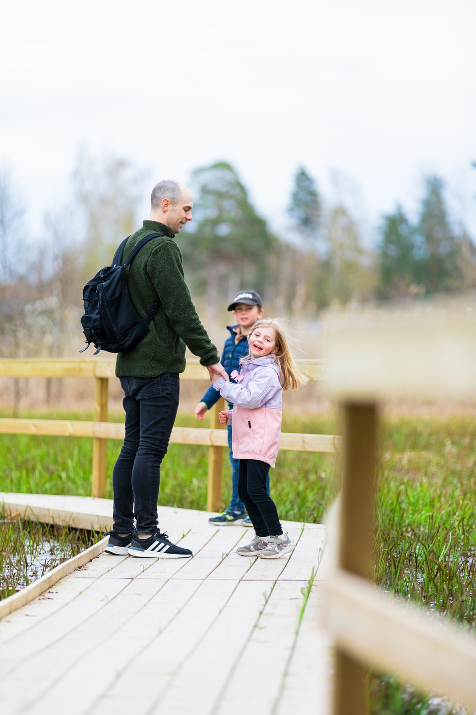 Tre personer, en förälder och två barn, vandrar på en vandringsled bestående av spångar. De håller varandra i händerna.