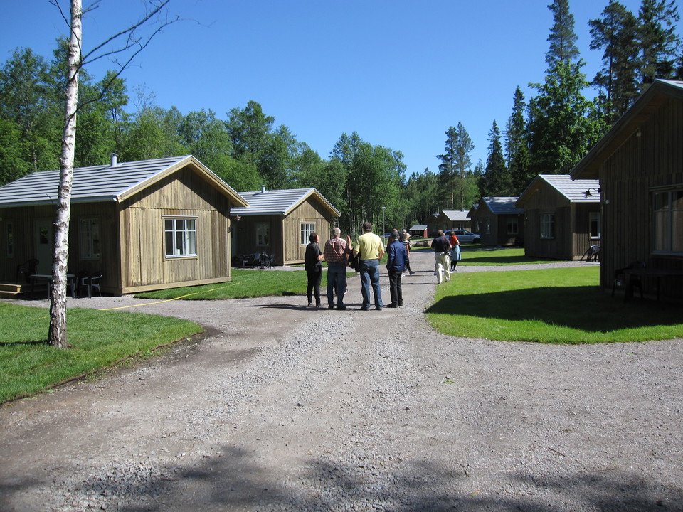 På en grusväg står en grupp människor. På båda sidorna av vägen står små trästugor på rad. 