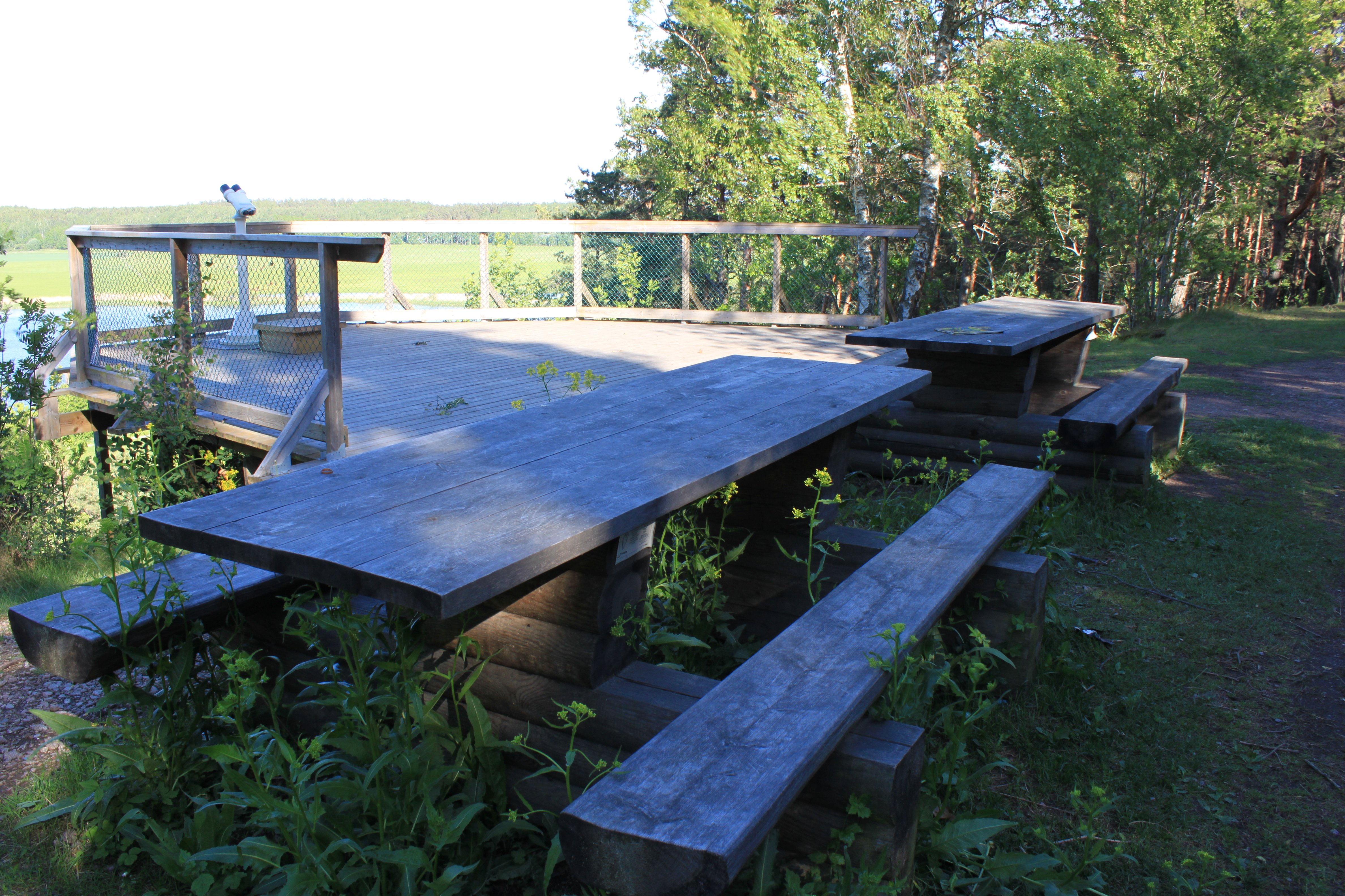 Två bänkbord står på gräsmark vid en rymlig utsiktsplattform med skyddsräcken runtom.