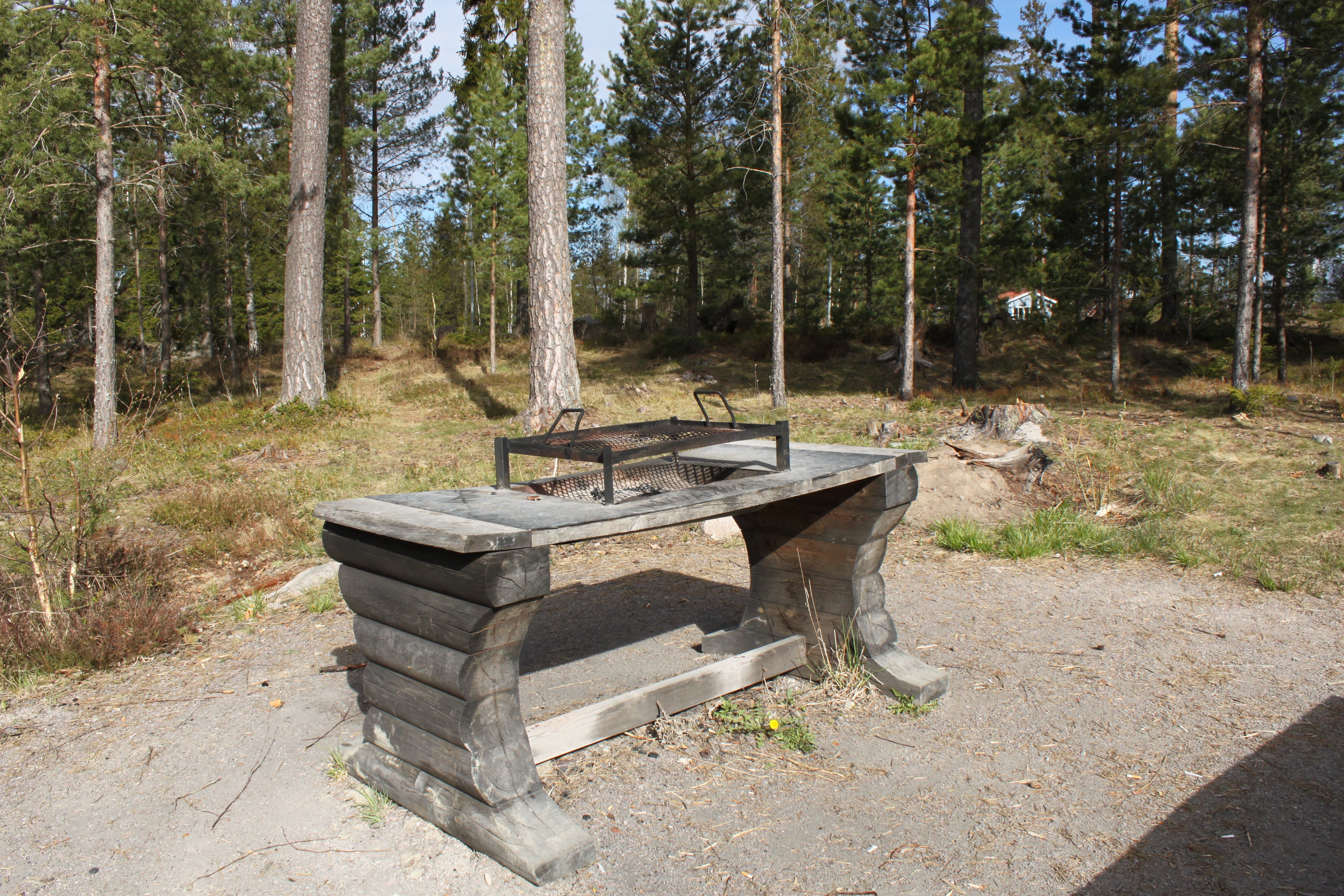 Ett grillbord med grillgaller står på en grusad yta med skog i bakgrunden.
