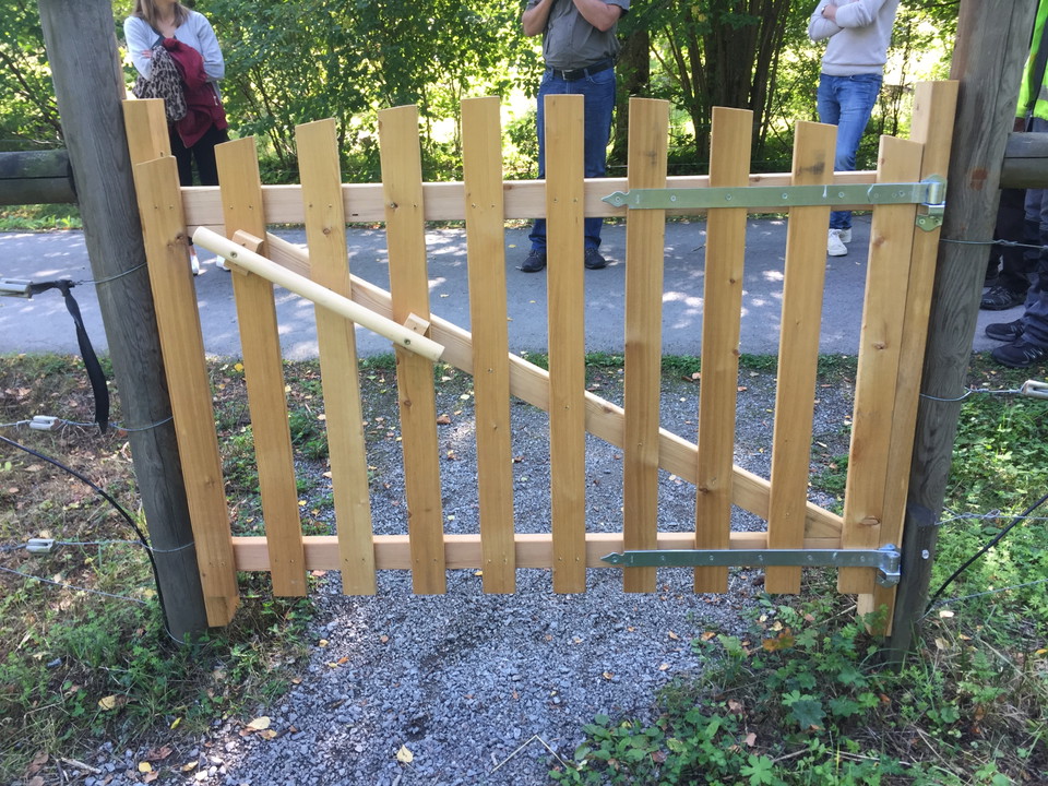 Närbild på en trägrind. Bakom grinden går en asfalterad stig och det står tre personer på stigen.