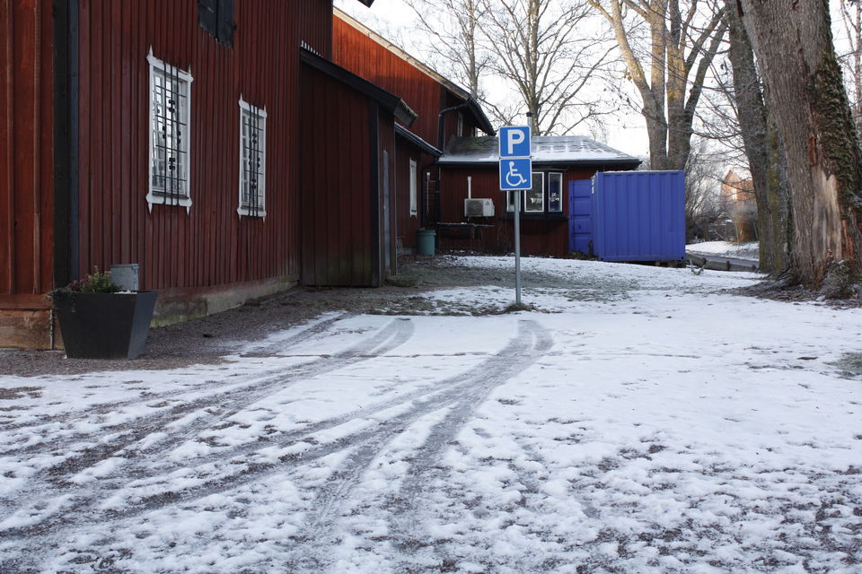 Vid en byggnad finns en öppen yta med skylt för handikapparkering. Det ligger snö på marken.