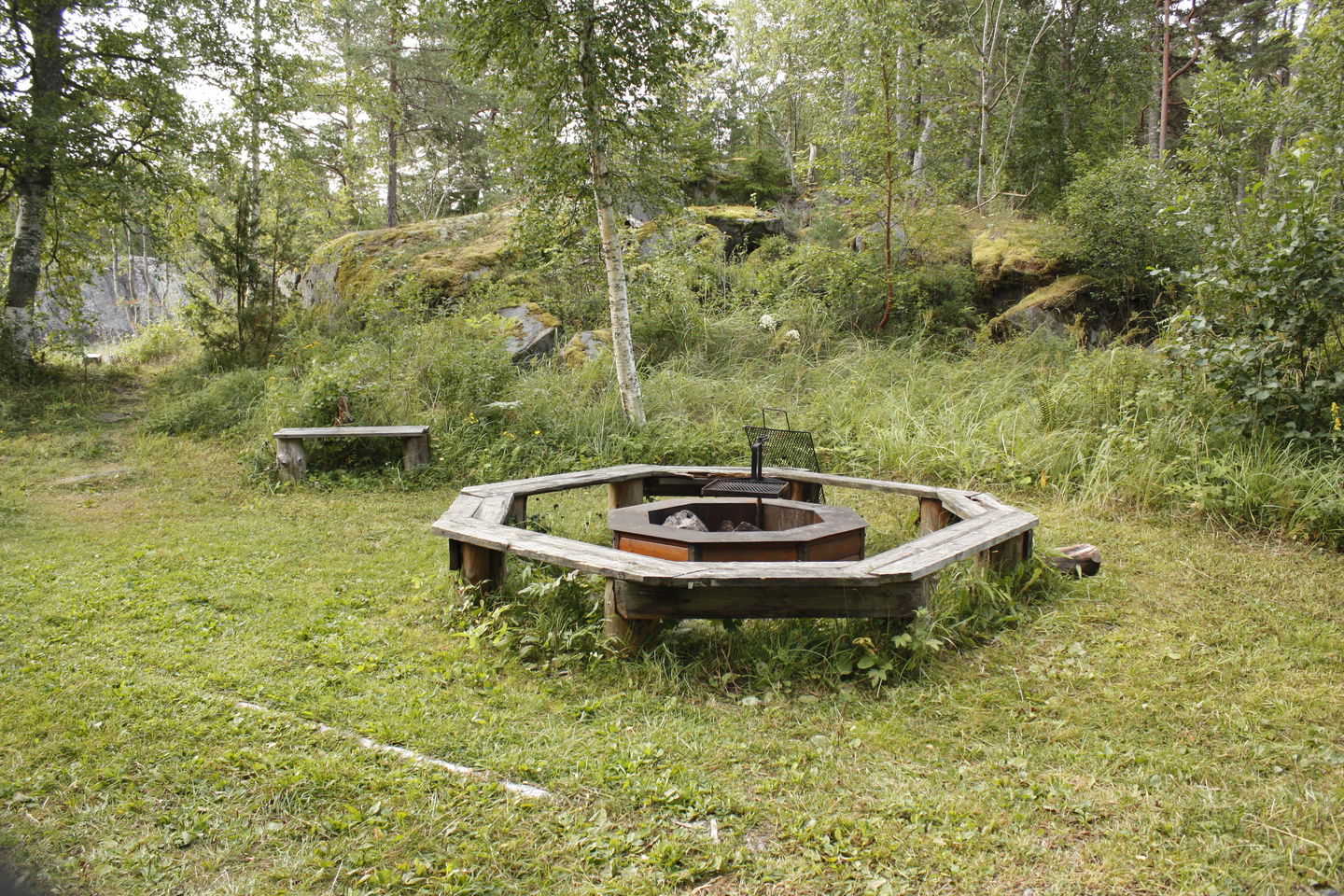 En grillplats med vridbart gallet står på gräsmark. Sittbänkar omringar grillplatsen.