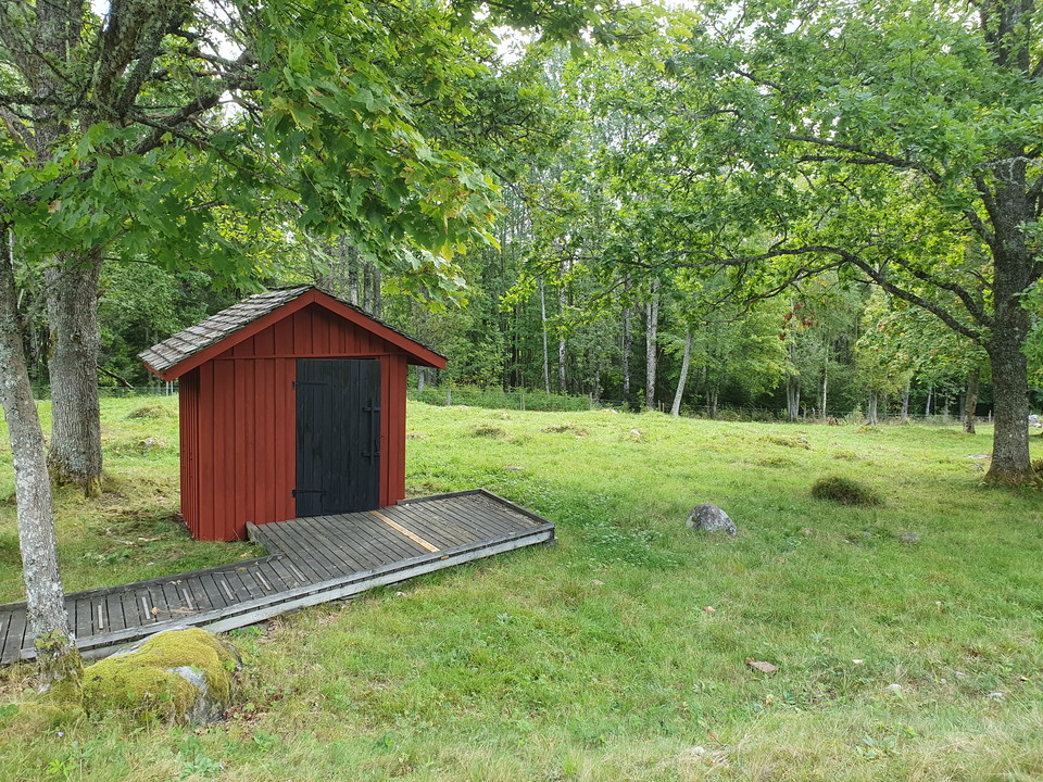 Torrtoalett med ramp som står i betesmark omgiven av lövträd.