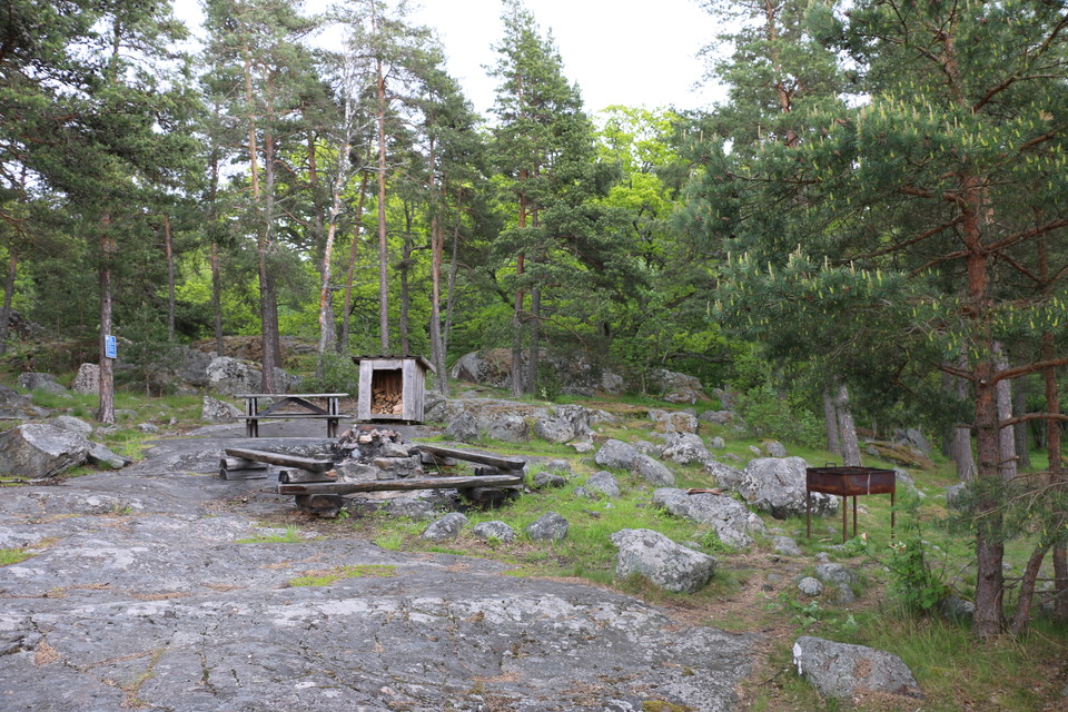 Rastplats med eldstad, vedförråd och bord och bänkar. Rastplatsen ligger på en klippa.