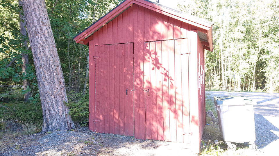 En torrtoalett med två dörrar står vid ett träd. Bredvid toaletter står en soptunna.