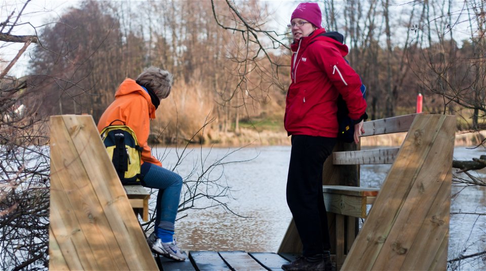 En mycket liten träplattform med två sittbänkar vid Fyrisån. På ena bänken sitter en person och blickar ut över vattnet och mittemot står en annan person och tittar mot kameran.