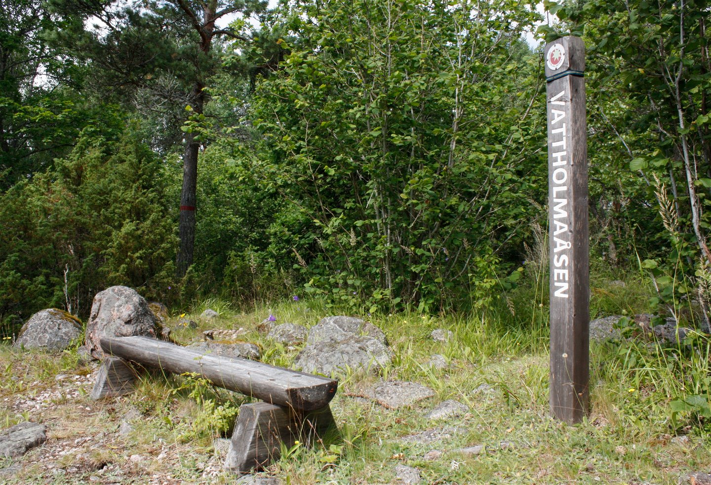 En sittbänk står på stenig yta i skogen. Vid bänken står en hög stolpe med texten Vattholmaåsen på.