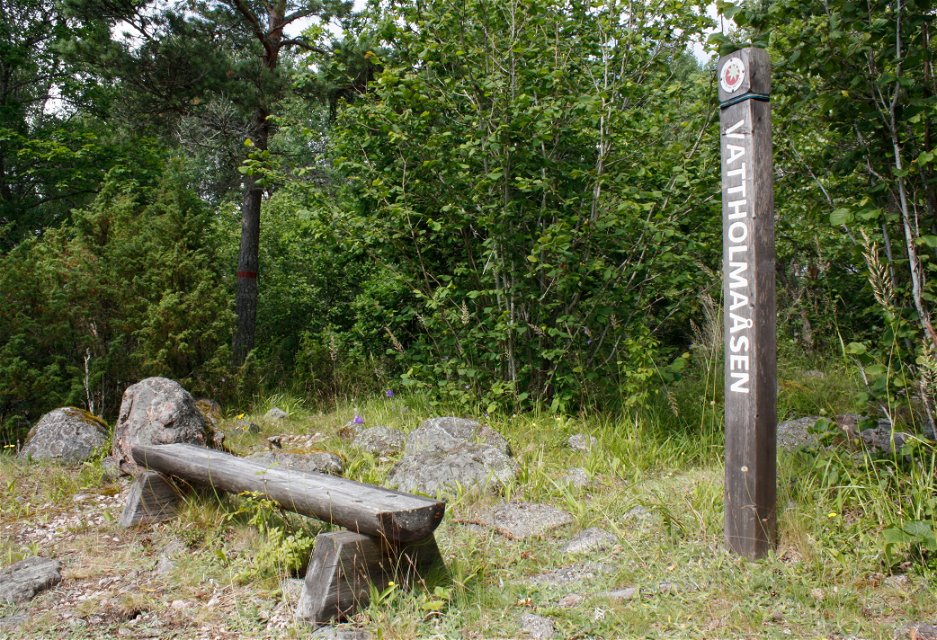 En sittbänk står på stenig yta i skogen. Vid bänken står en hög stolpe med texten Vattholmaåsen på.