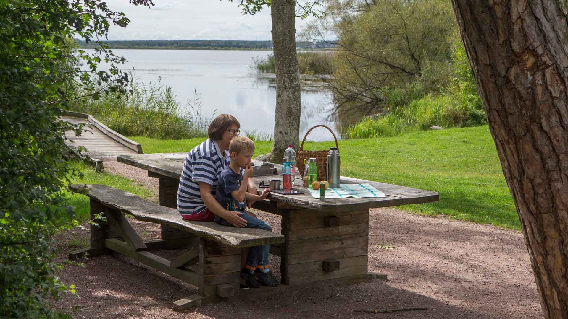 En kvinna och ett barn sitter med uppdukad fika vid ett bänkbord med utsikt över sjön