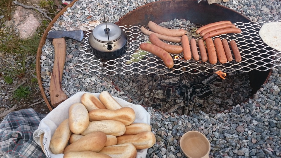 Eldstad med korv och en kaffepanna på ett galler över elden. Vid sidan ligger en yxa och en korg full av korvbröd.