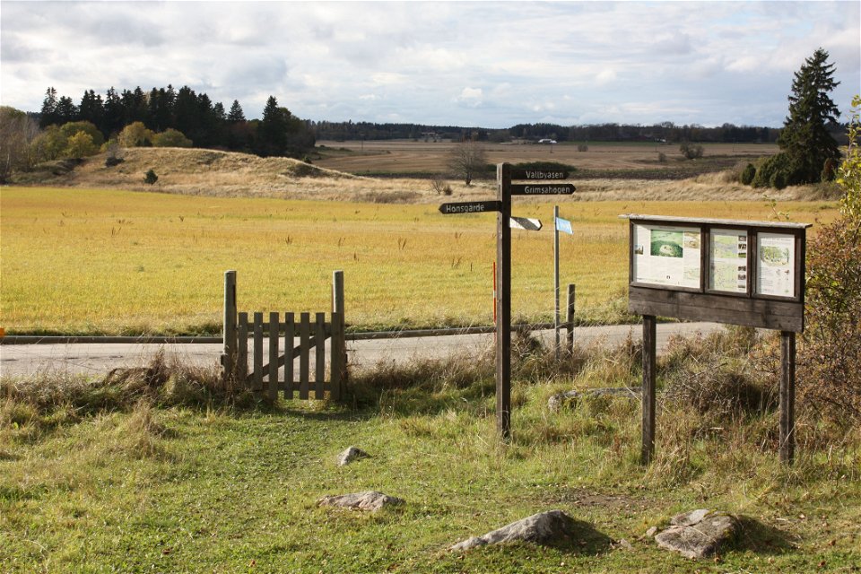 På ena sidan av en mindre bilväg står en grind. Innanför grinden finns en informationstavla och en stolpe med pilar som pekar i olika riktningar. Marken är ojämn med gräs och flera större stenar.