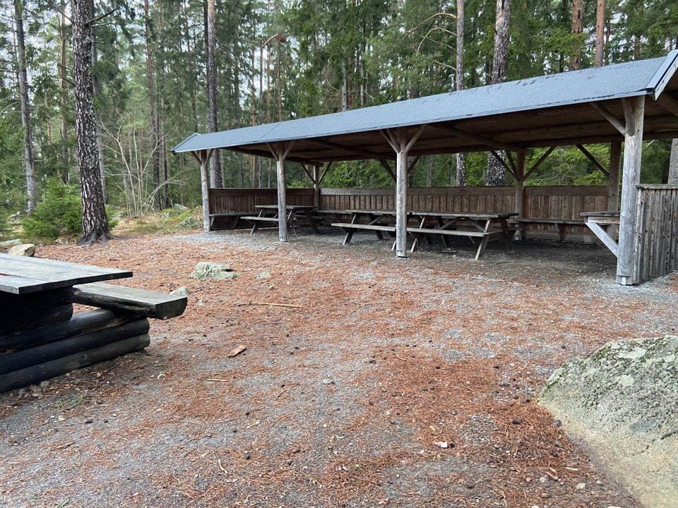 En långsträckt byggnad som består av tak, tre väggar och en öppen sida. Under taket står några bänkbord.