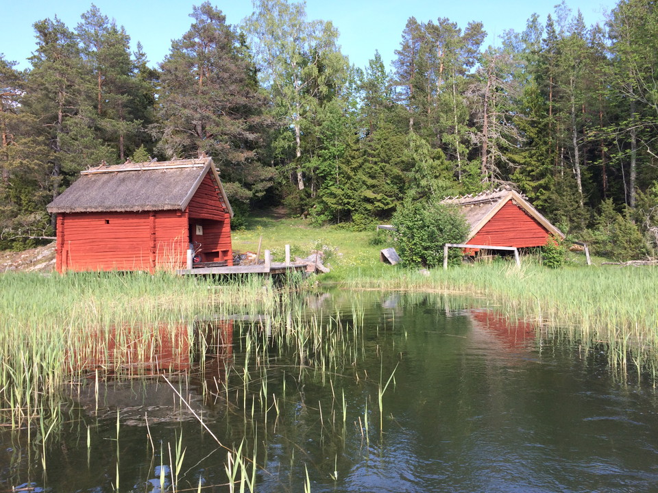 Vy från vattnet mot sjöbod, brygga och båthus. Skog i bakgrunden.
