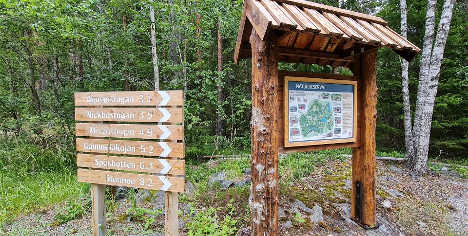 Till höger står en stigvisare med 6 destinationer i Florarna. Till vänster står en informationstavla som sitter på ett ställ med tak. I bakgrunden björkar och gröna löv.