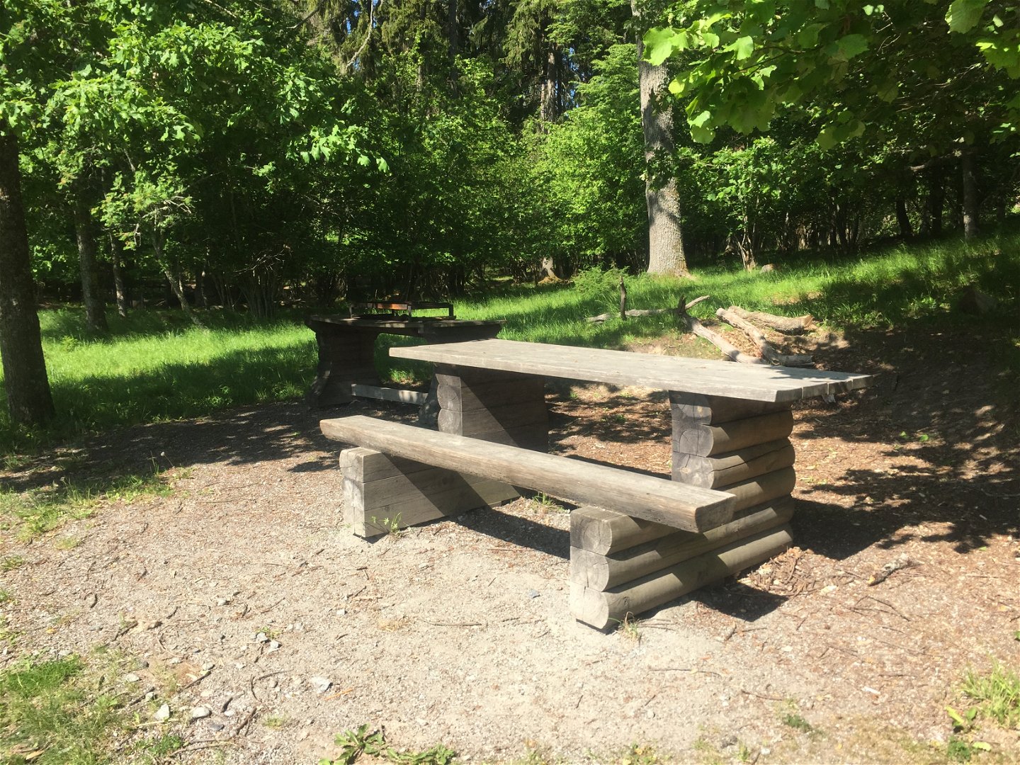 Ett bord med bänk på ena sidan står på en grusyta i skogen. Bredvid står ett grillbord.
