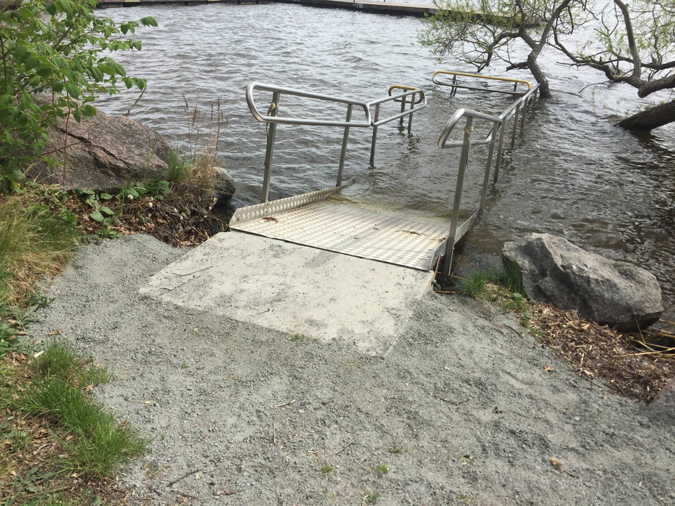 En ramp med handräcken leder ner i vattnet. Framför rampen är det grus och en jämn betongplatta.