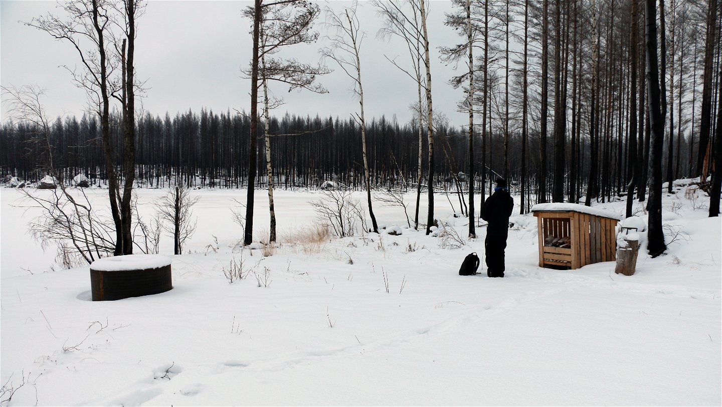 En svartklädd person står vid grillplats och vedförråd vid en snötäckt sjö.