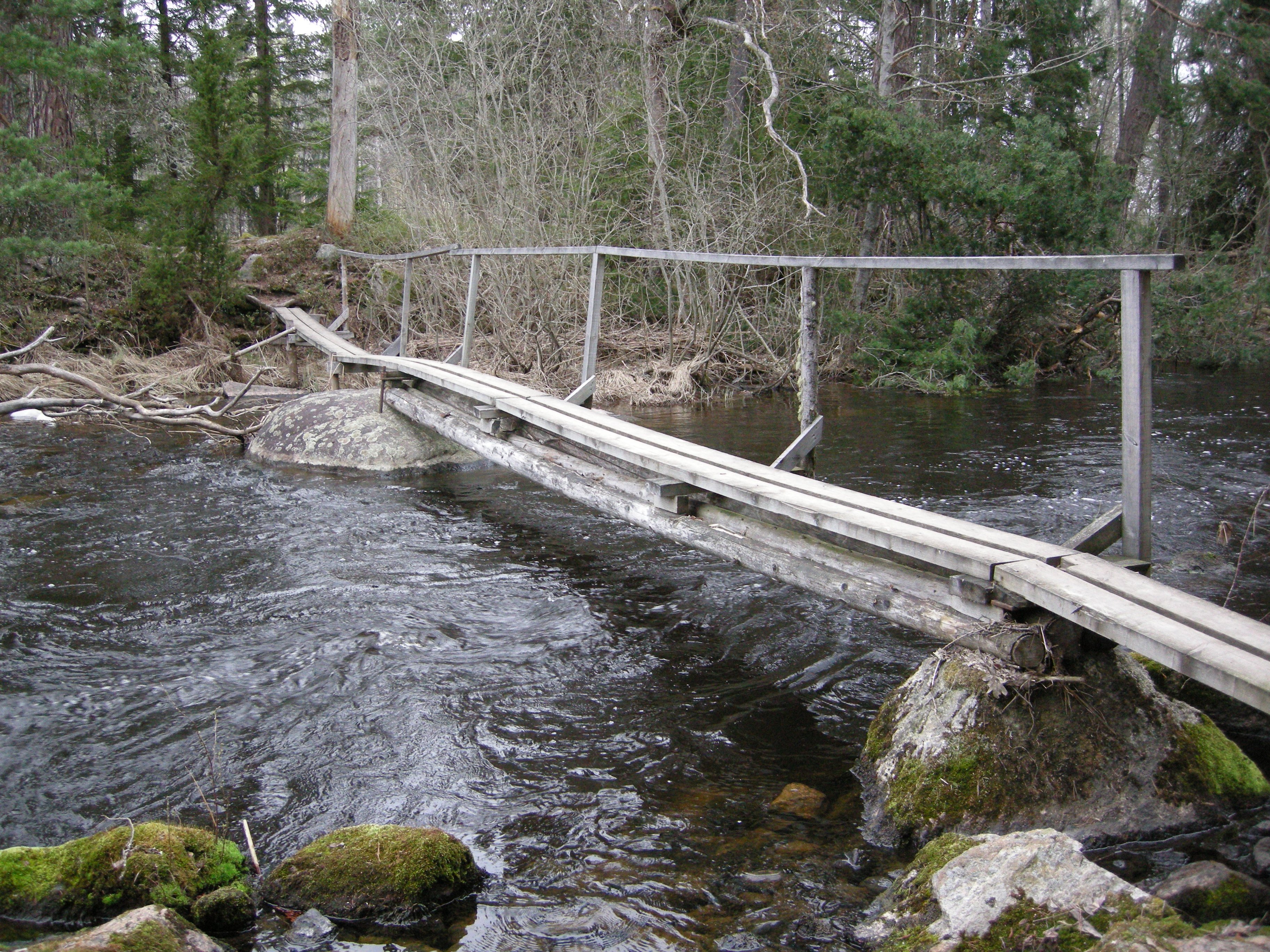 En mycket smal bro med ett handräcke på ena sidan går över ett vattendrag.