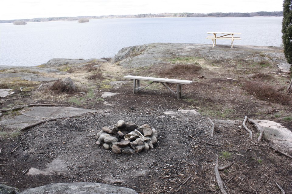 En eldstad av sten uppe på stenhäll vid vattnet. Längre bort finns en sittbänk och ett bänkbord.
