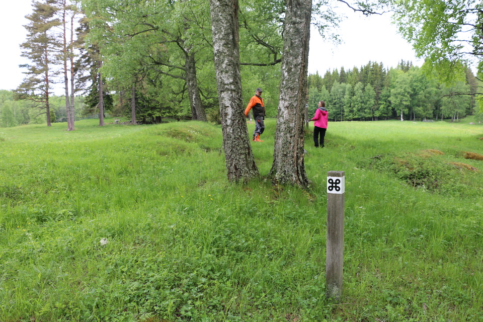 Vitmålad stolpe med svart kringla. Två personer går bortåt på ängsmark.