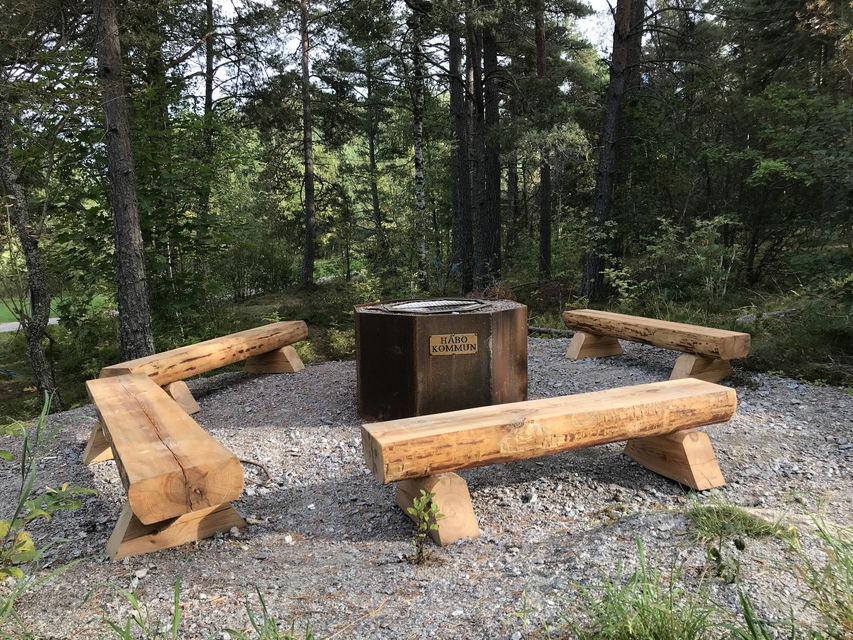 Fyra stockbänkar står på en grusad yta runt en eldstad.