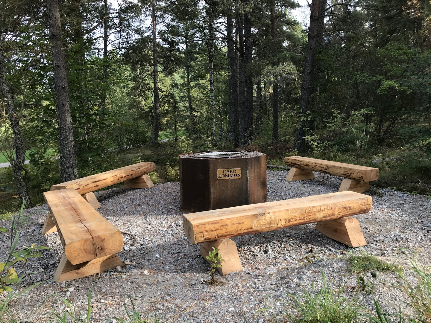 Fyra stockbänkar står på en grusad yta runt en eldstad.