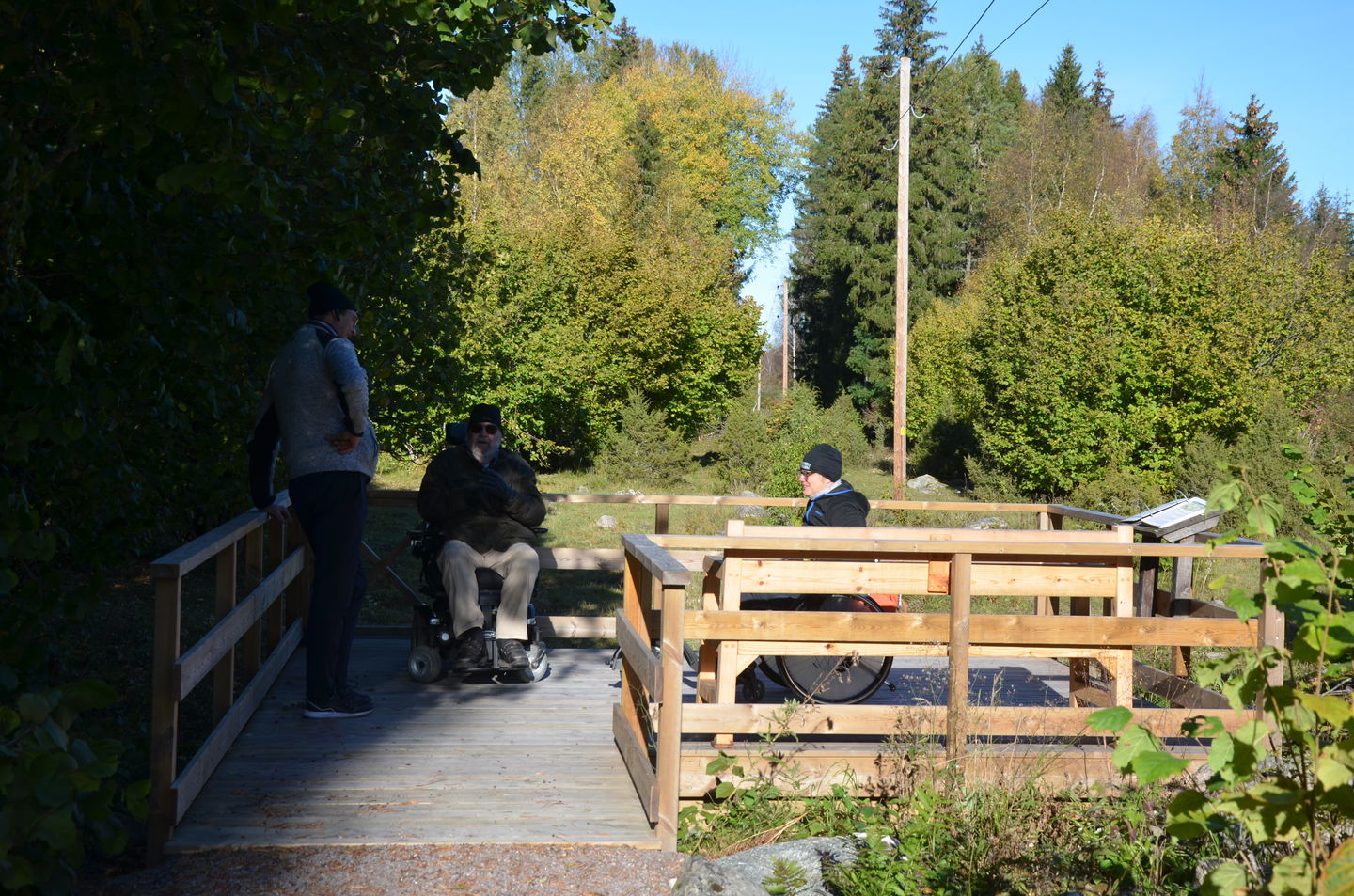 En byggd trall med staket runt. 2 personer i rullstol och en stående på rastplatsen med lummig grönska runt