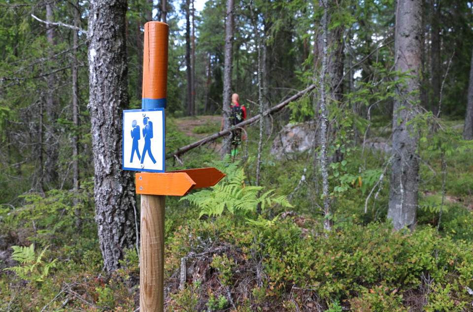 Stolpe med orange pilar som visar vägen. På stolpen sitter även en skylt som symboliserar två gående personer.